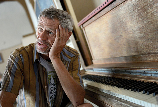 Image shows a man with tanned skin and grey hair looking into the distance while he rests his head on his hand and is sitting in front of a piano.